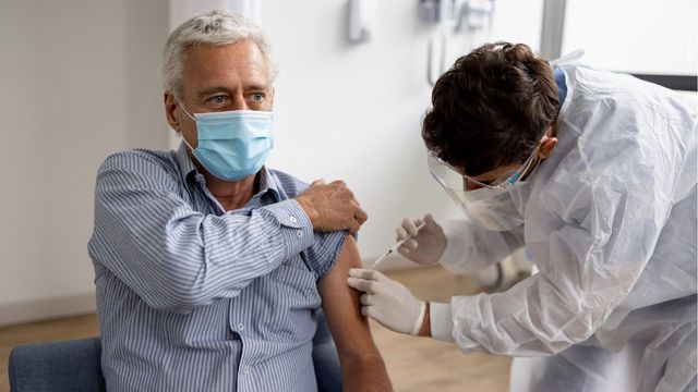 A man wearing a mask receiving a vaccine in his left arm. 