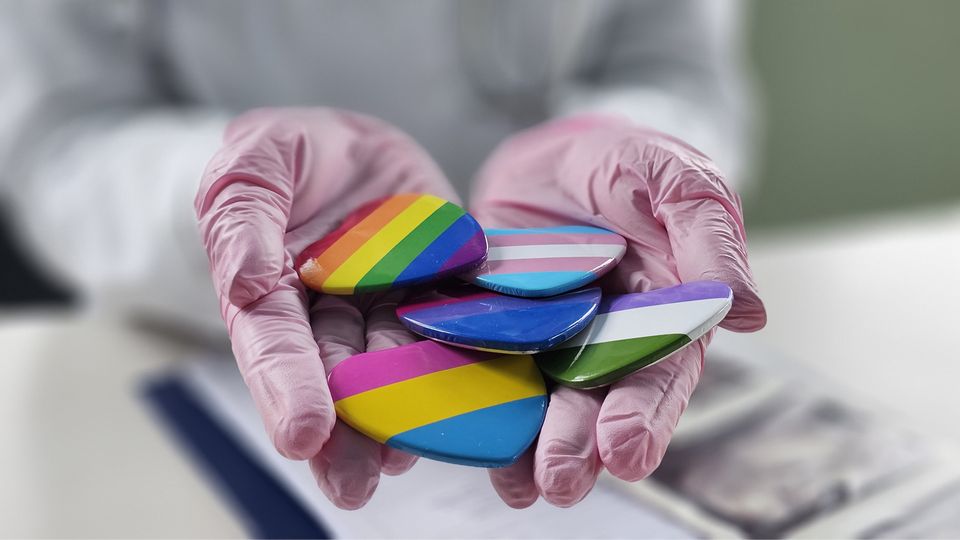 The gloved hands of a scientist holding badges displaying various LGBTQIA+ pride flags.