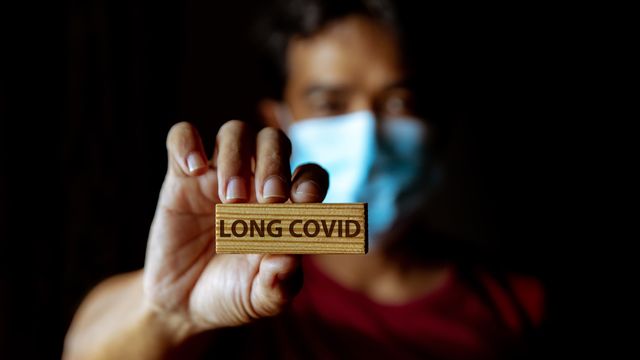 Person wearing a face mask holding a wooden block engraved with the words "LONG COVID." 