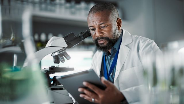 Scientist in a lab coat analyzing data on a tablet beside a microscope in a research laboratory. 