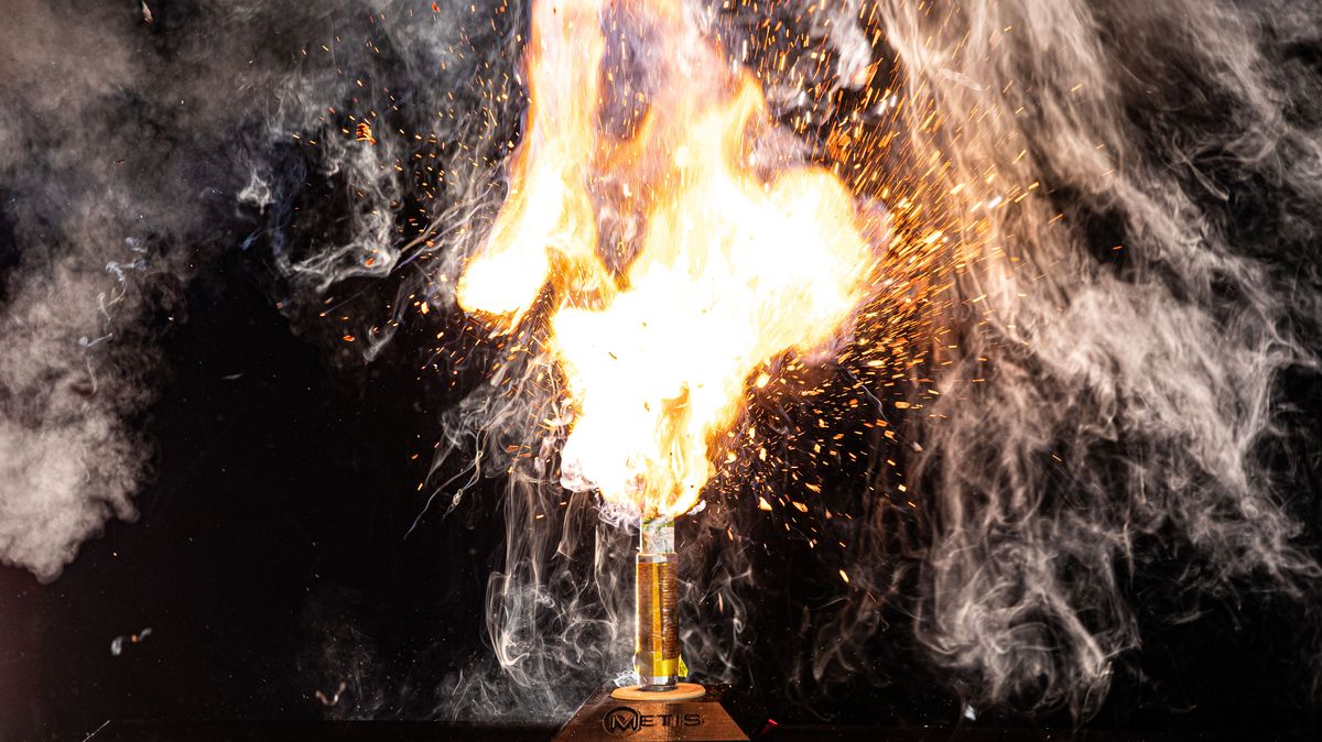 A battery on a small metal plinth. The battery is emitting flames and smoke upwards as it experiences a thermal runaway event.