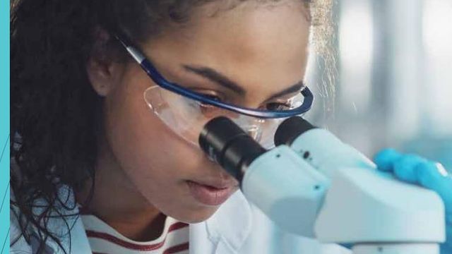 A scientist wearing lab goggles looking through a microscope 