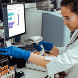 A scientist in a lab coat and gloves scans a sample tube with a barcode scanner, while a computer screen displays lab data. 