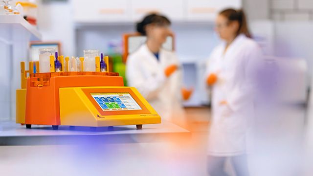 Image of a orange and yellow lab instrument with a digital touchscreen and test tubes on top is prominently shown on a workbench, with two scientists in white lab coats. 
