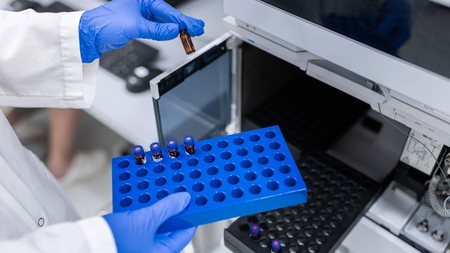 A person wearing blue gloves places small vials into a blue sample holder in a laboratory, with scientific equipment visible in the background. 