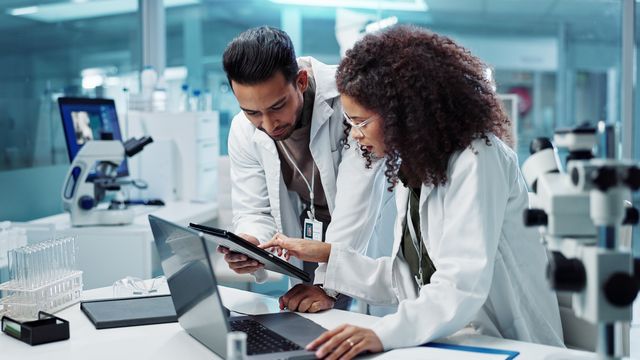 Two researchers in lab coats reviewing data on a tablet and laptop in a modern laboratory. 