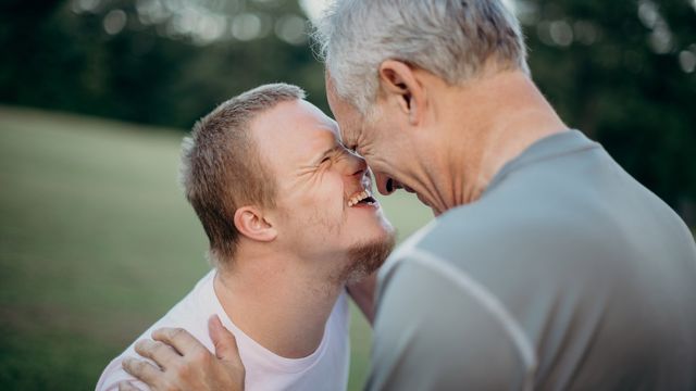 Older man and young adult with Down syndrome laughing and touching foreheads outdoors. 